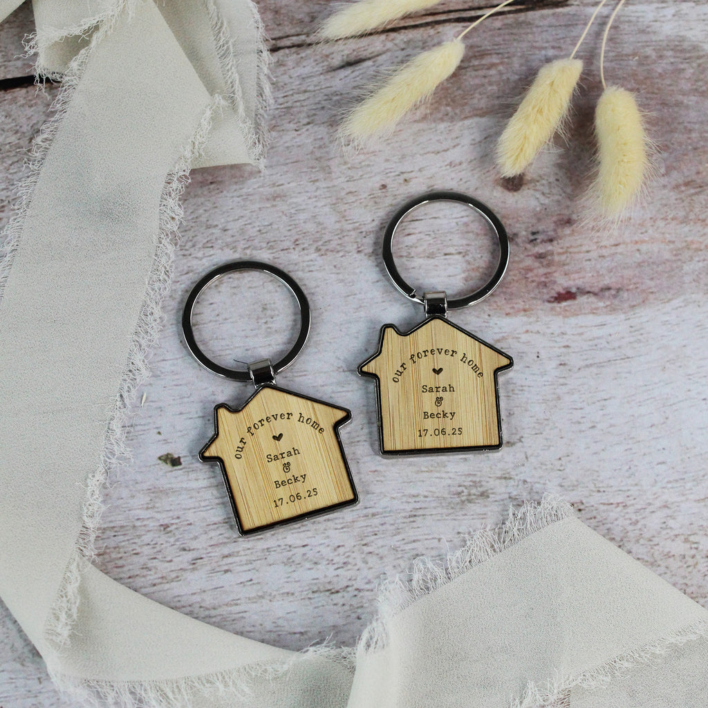Two wooden keychains shaped like houses on a textured surface with ribbons and dried plants.
