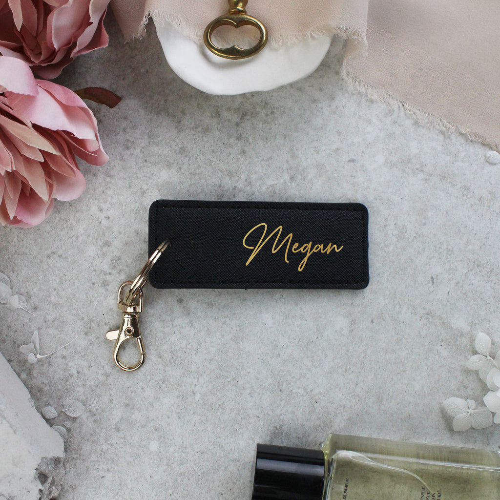 Black keychain with 'Megan' engraving on a gray surface with flowers and a bottle.