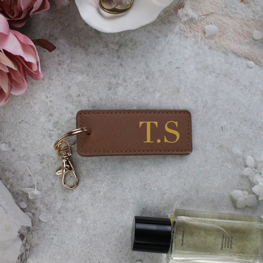 Brown leather keychain with 'T.S' initials on a gray surface with flowers and a bottle.
