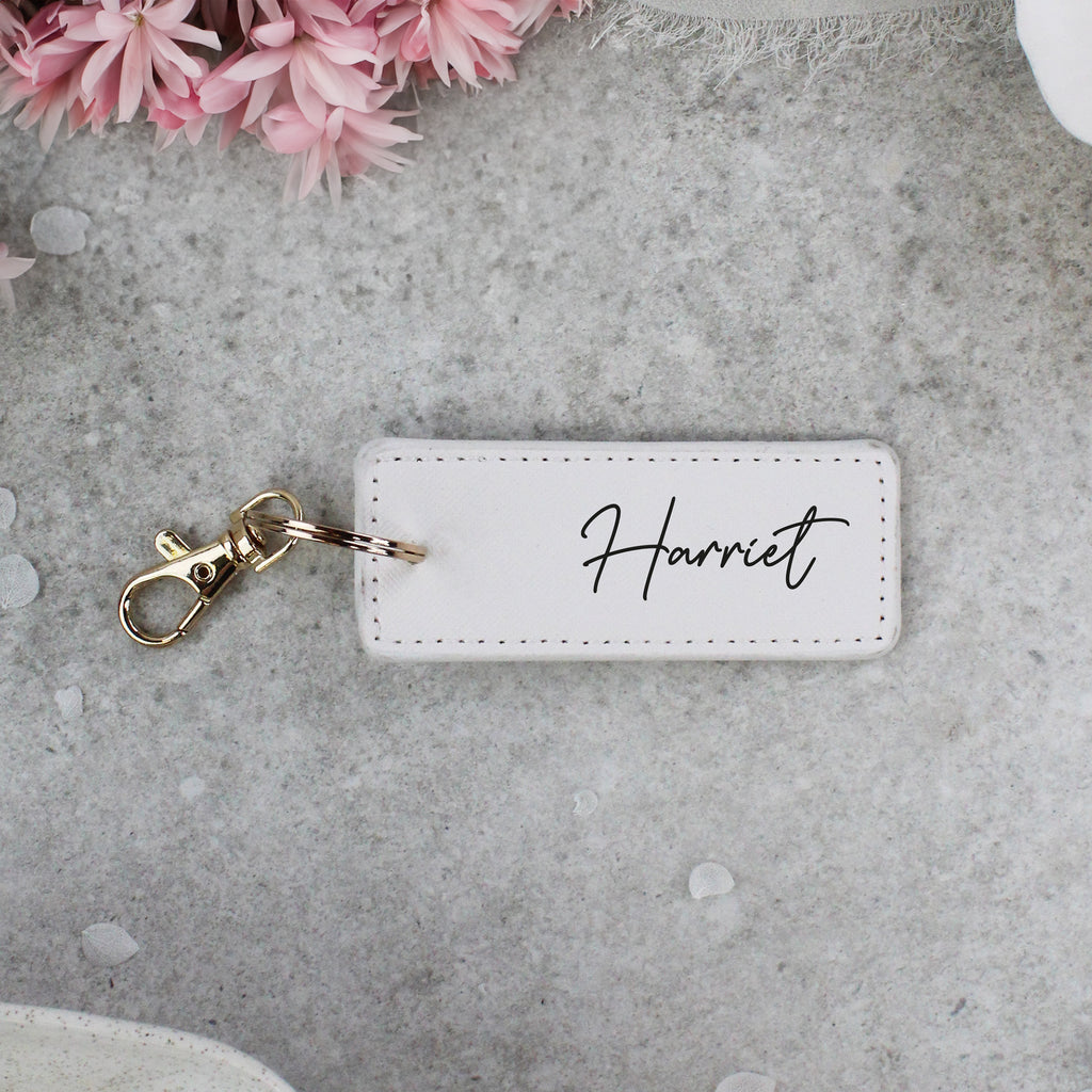 White keychain tag with 'Harriet' engraved, on a gray surface with pink flowers.