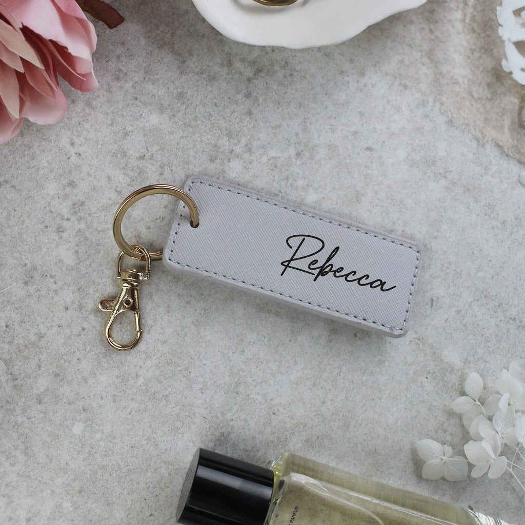 Keychain with 'Rebecca' tag on a textured surface with flowers and a bottle.