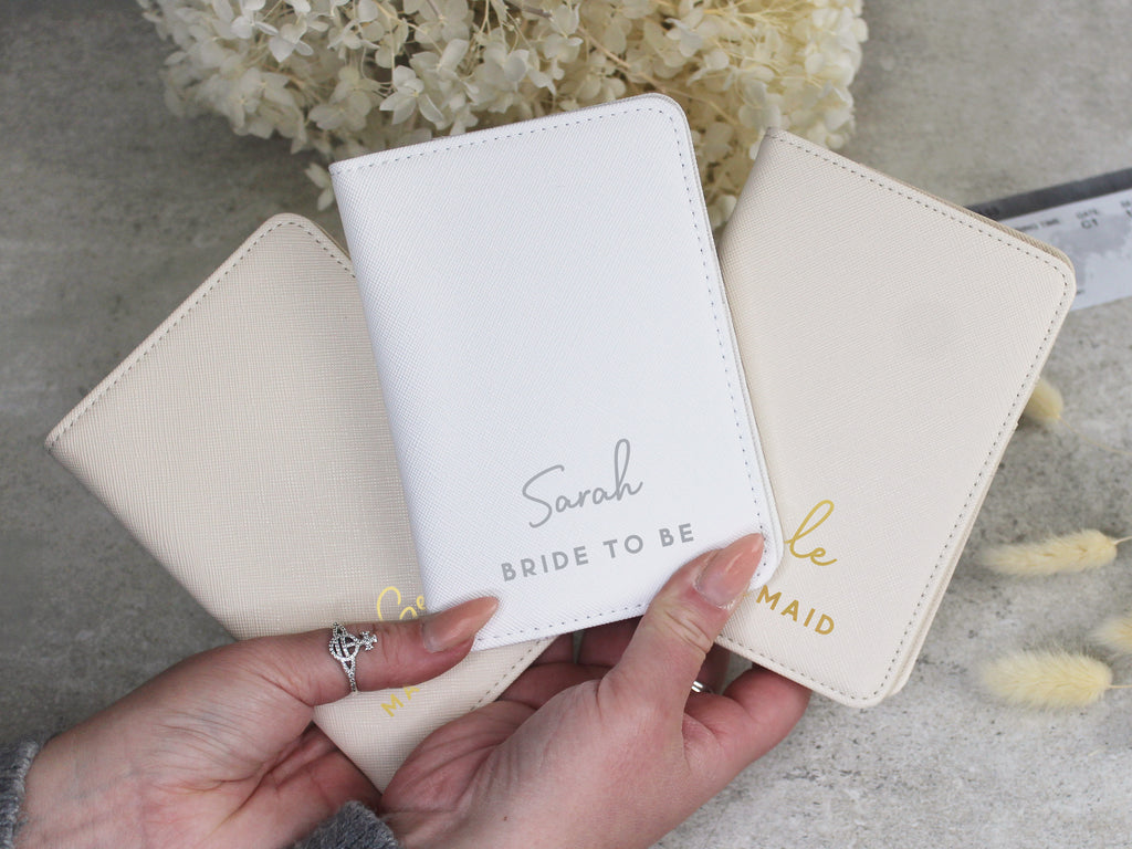 Three white passport holders with personalized text held by a hand, with flowers in the background.
