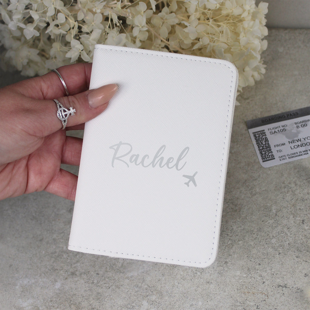 White passport holder with 'Rachel' engraved, held by a hand with rings, on a light surface with flowers in the background.