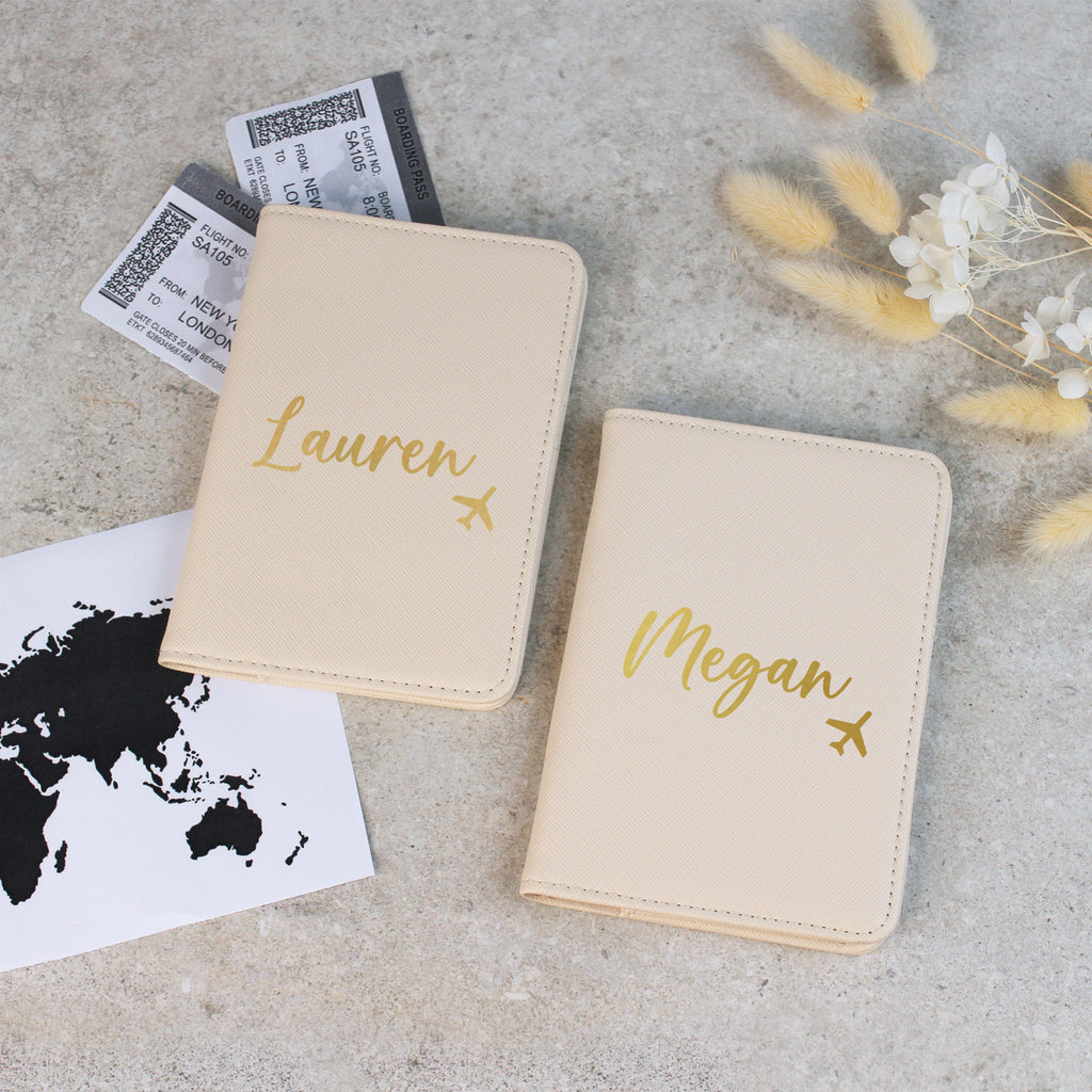 Two beige passport holders with 'Lauren' and 'Megan' printed on them, surrounded by travel-related items.