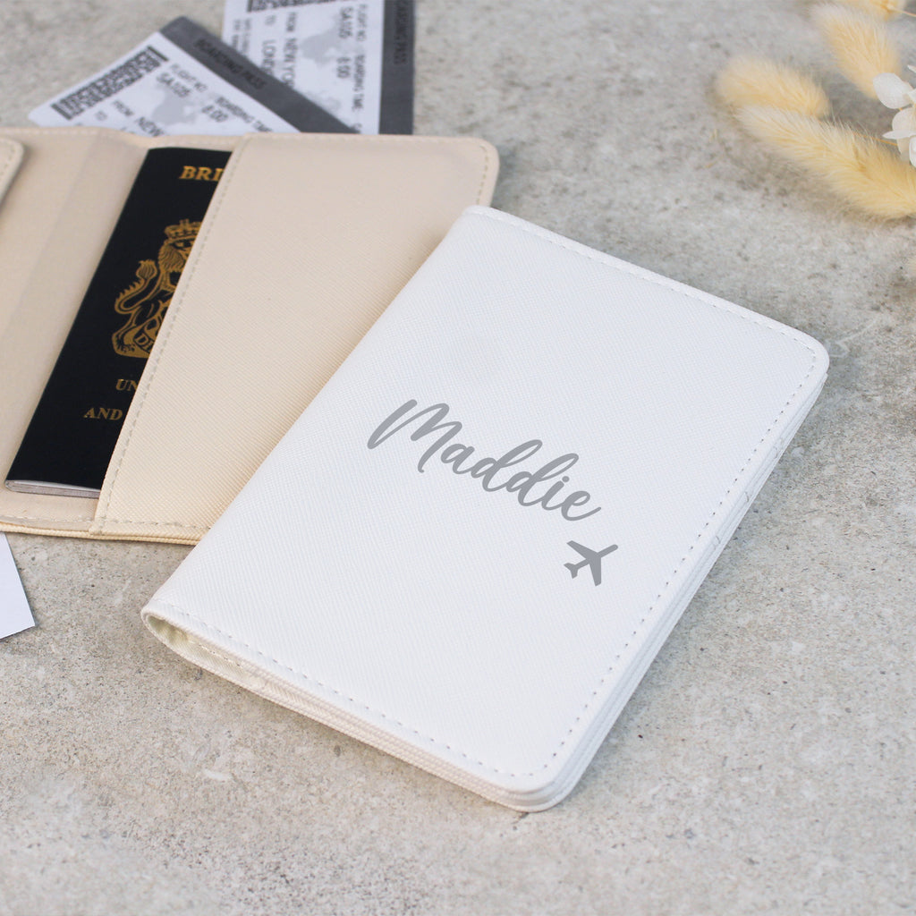 White passport holder with 'Maddie' and an airplane icon on a concrete surface with passports and tickets.