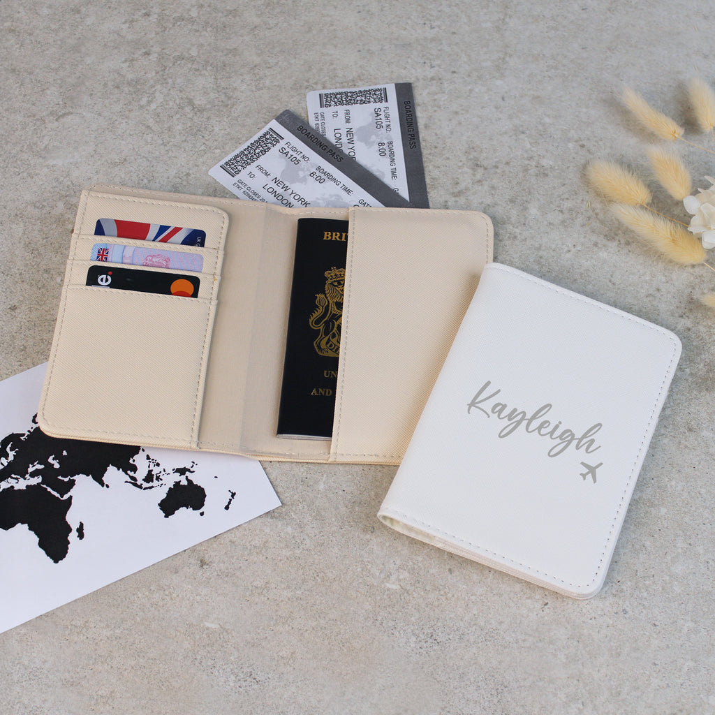 Beige passport holder with cards and a passport, next to a white notebook with 'Kayleigh' on a gray surface.