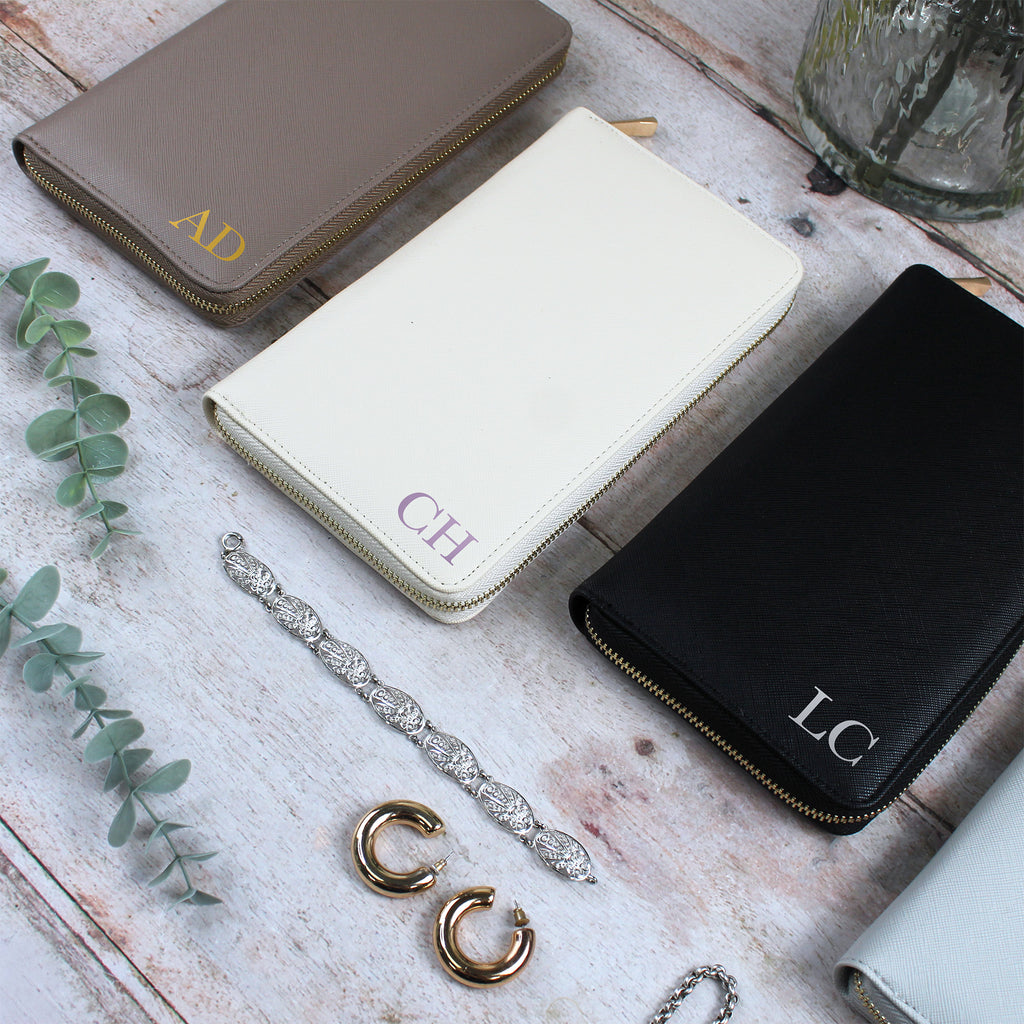 Three monogrammed wallets on a wooden surface with jewelry and a plant.