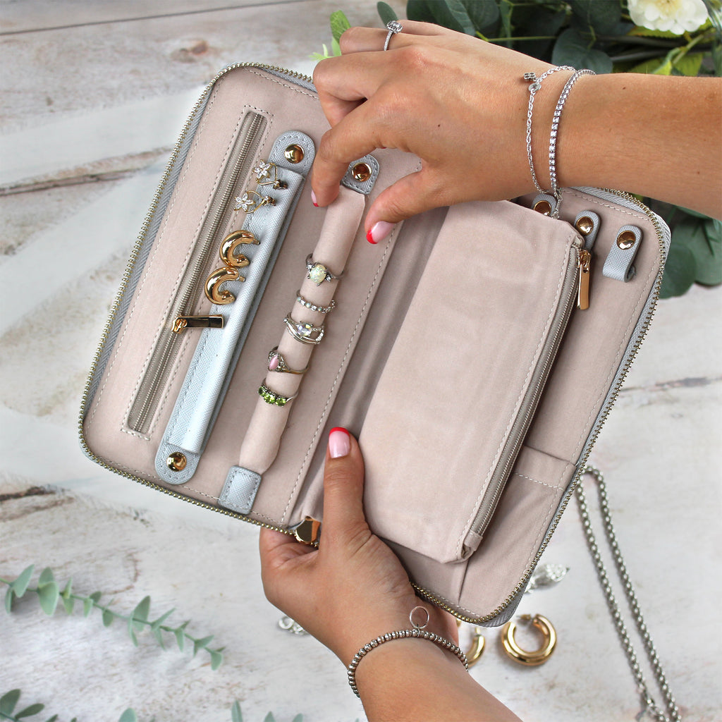 Hand holding a beige clutch with jewelry displayed on a light wooden surface.