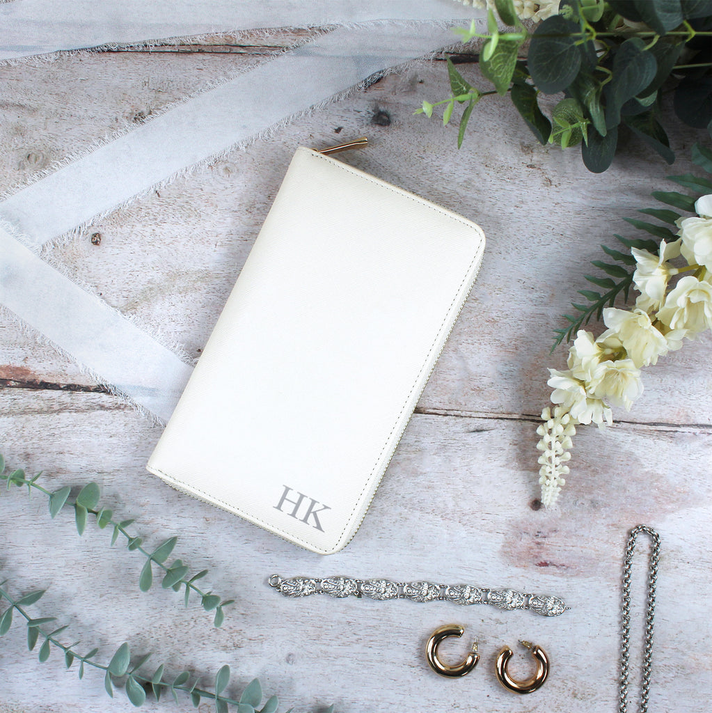 White leather wallet with initials, jewelry, and flowers on a wooden surface
