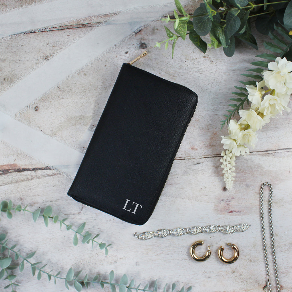 Black jewelry pouch with initials, gold hoop earrings, and a necklace on a wooden surface with flowers.