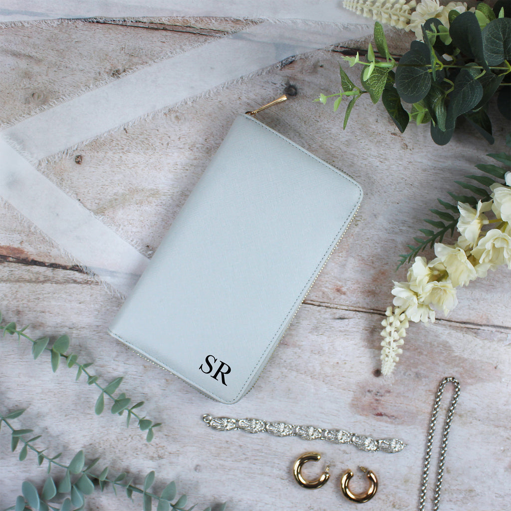 Light blue wallet with 'SR' initials, gold earrings, bracelet, and flowers on a wooden surface