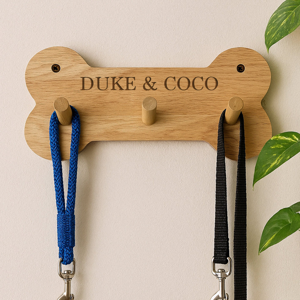 Wooden dog bone-shaped leash holder with 'DUKE & COCO' engraving, holding two leashes against a light background.
