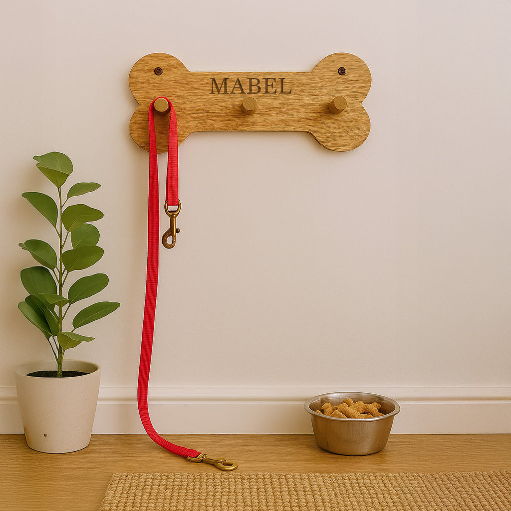 Wooden bone-shaped leash holder with 'MABEL' engraved, red leash, and metal dog bowl on a wooden floor.