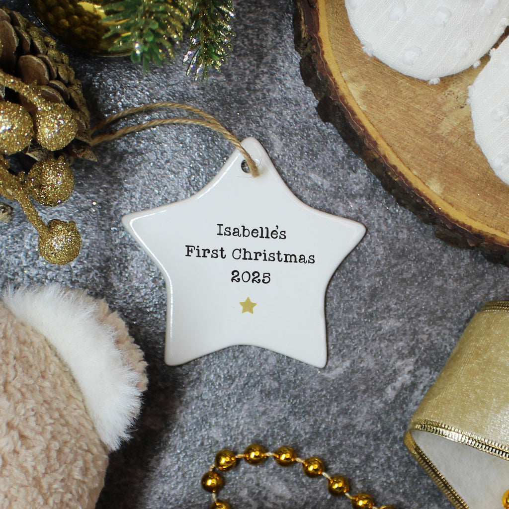 Star-shaped ornament with 'Isabellés First Christmas 2023' text, surrounded by festive decorations on a gray surface.