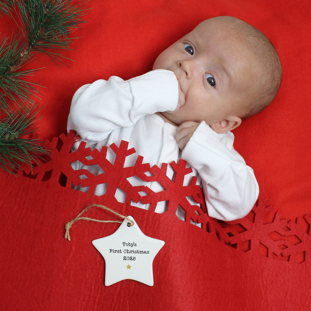 Baby lying on a red blanket with Christmas decorations, including a snowflake and a 'First Christmas' tag.