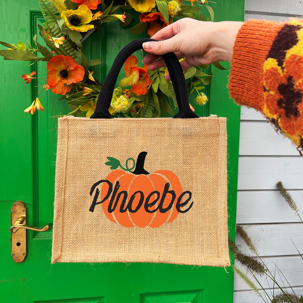 Person holding a burlap bag with a pumpkin design and 'Phoebe' text in front of a green door with a wreath.