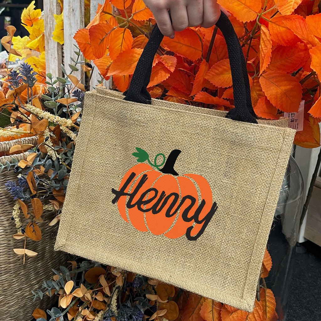 Jute bag with pumpkin design and 'Henny' text held by a hand against an autumn leaf background.