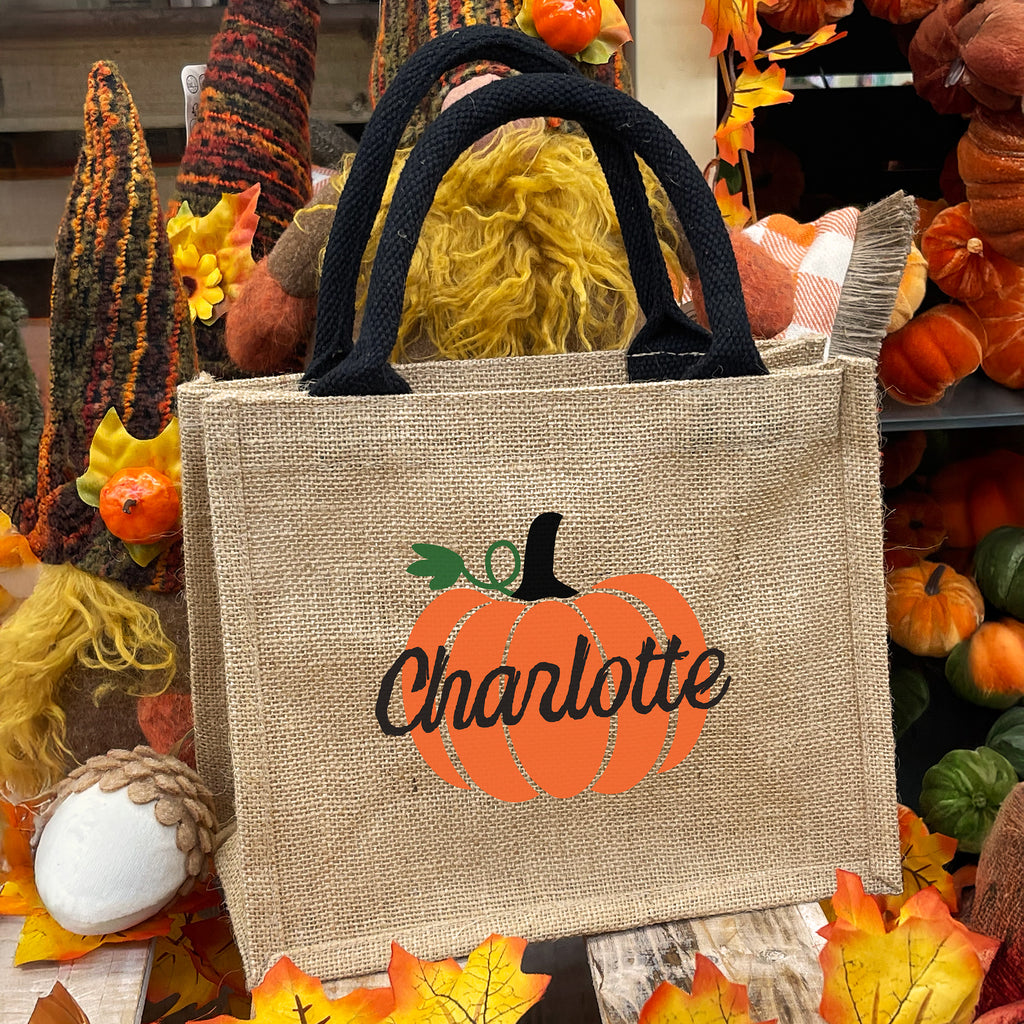Jute bag with pumpkin design and 'Charlotte' text, surrounded by autumn decorations.