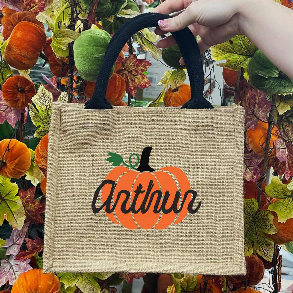 Burlap bag with a pumpkin design and 'Arthur' text, held against a autumn-themed background.