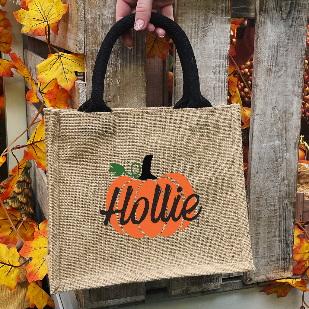 Jute bag with pumpkin design and 'Hollie' text, held by a hand against a wooden background with autumn leaves.