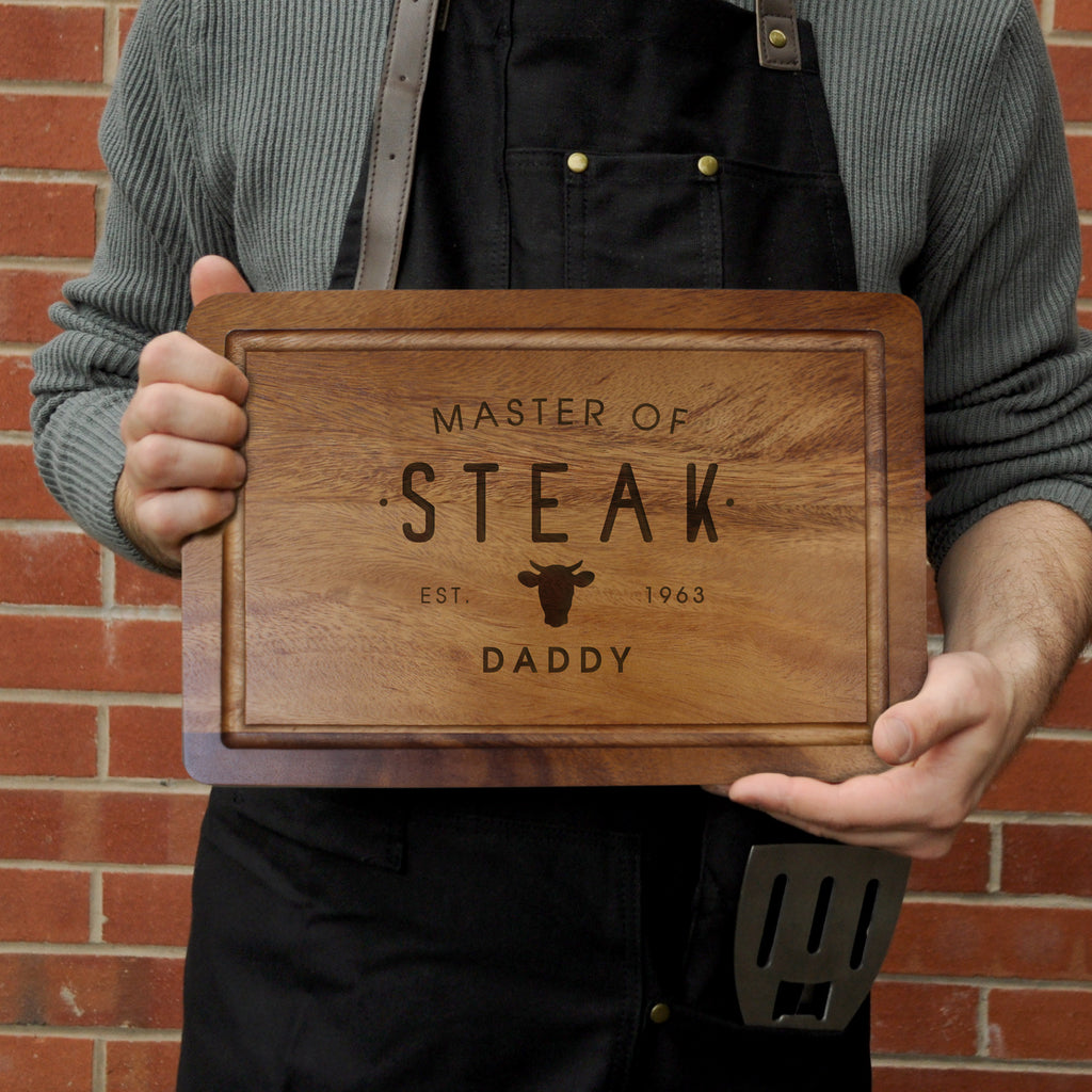 Person holding a wooden cutting board with 'Master of Steak' engraving against a brick wall.