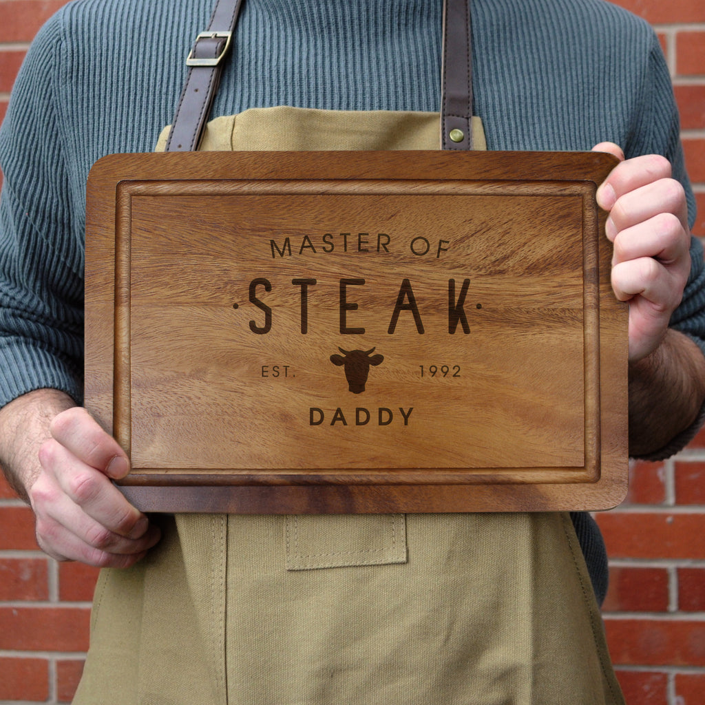 Person holding a wooden cutting board with 'Master of Steak' engraving against a brick wall.