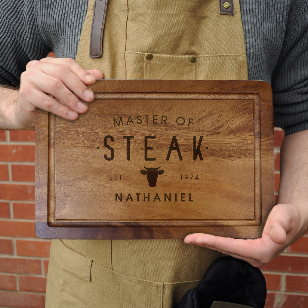 Person holding a wooden cutting board with 'Master of Steak' engraving