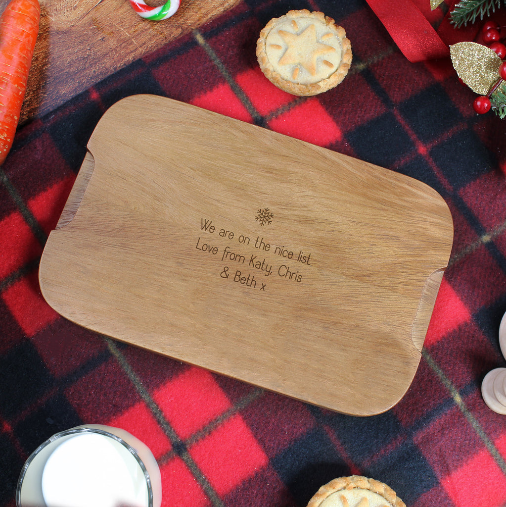 Wooden cutting board with engraved message on a red plaid tablecloth with Christmas decorations.