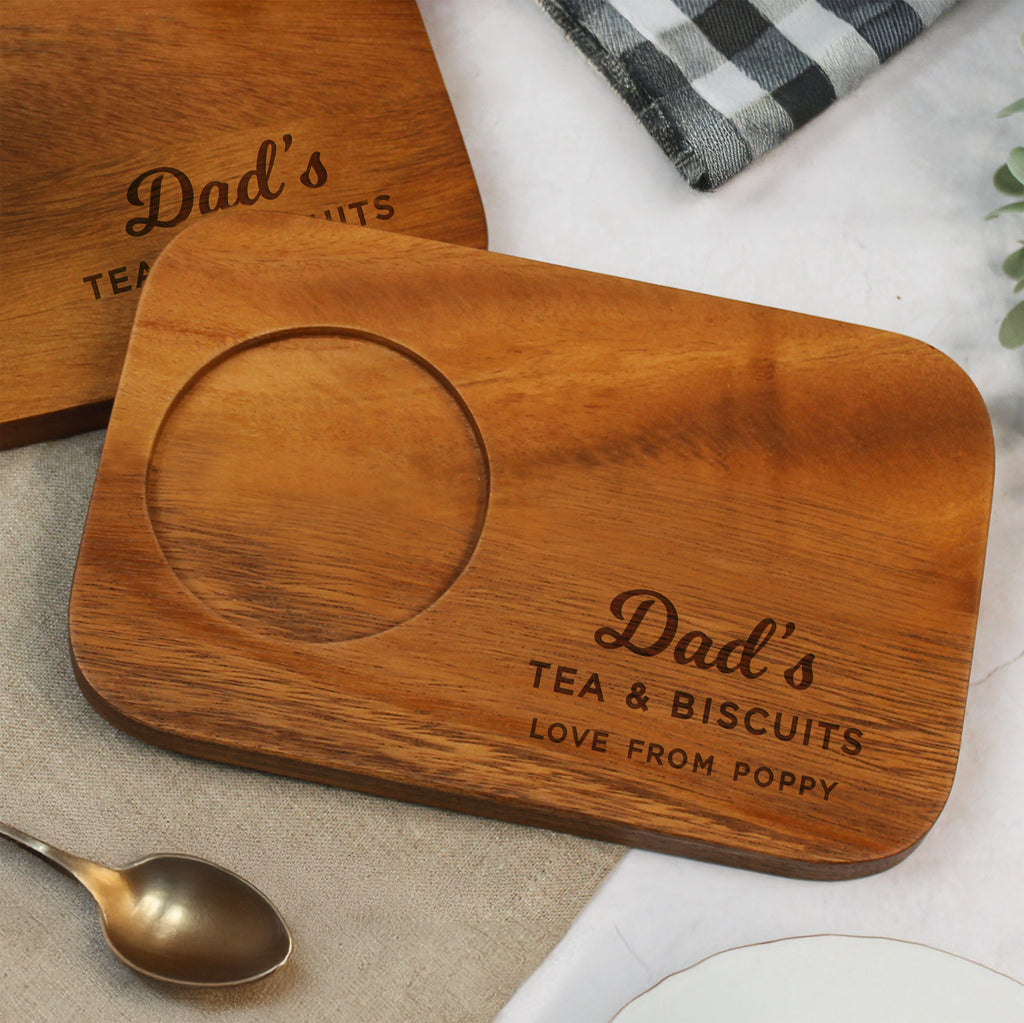 Wooden tray with 'Dad's Tea & Biscuits' engraving on a table with a spoon and plaid cloth.