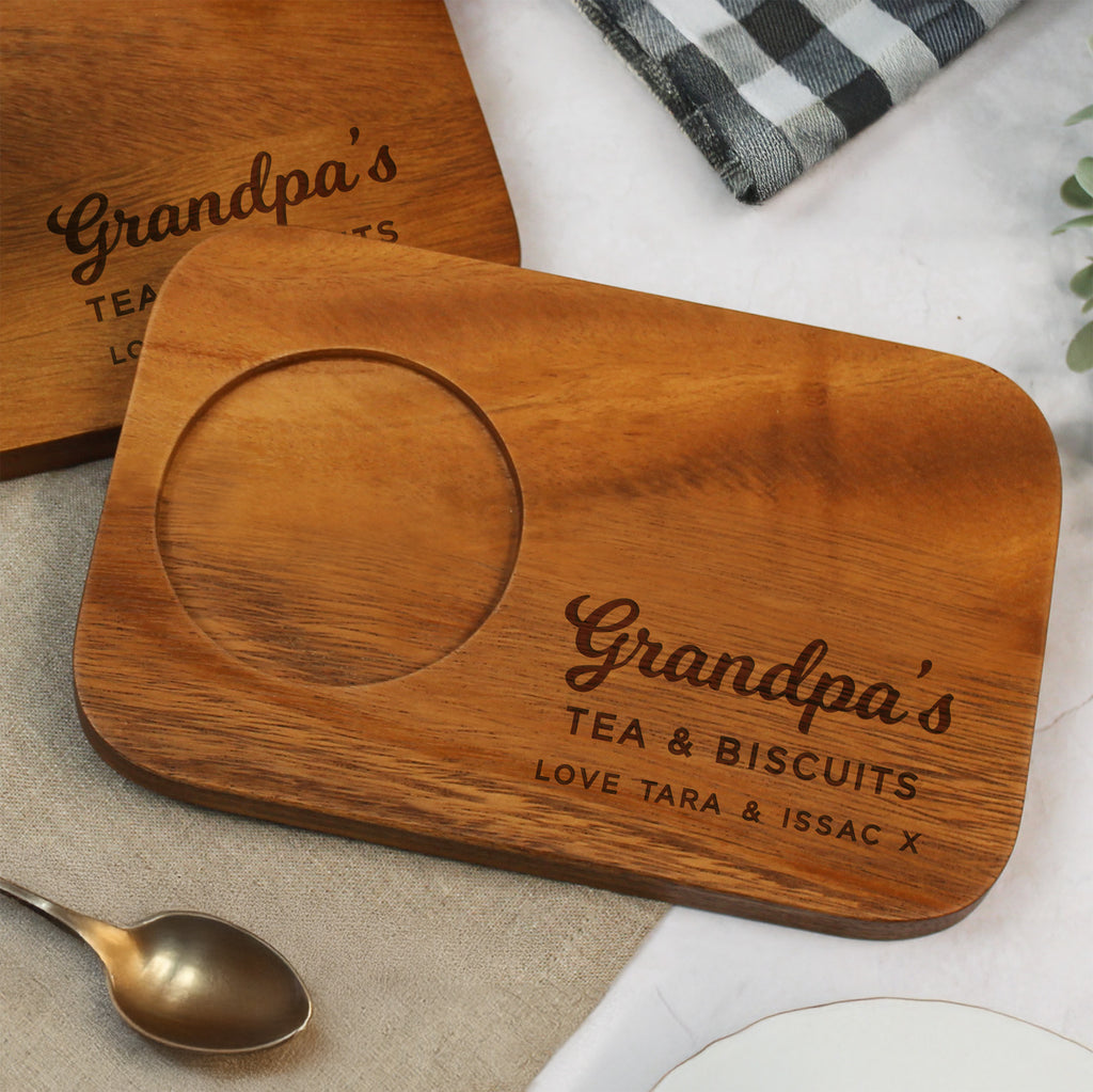 Wooden tray with 'Grandpa's Tea & Biscuits' engraving on a table with a spoon and plaid cloth.