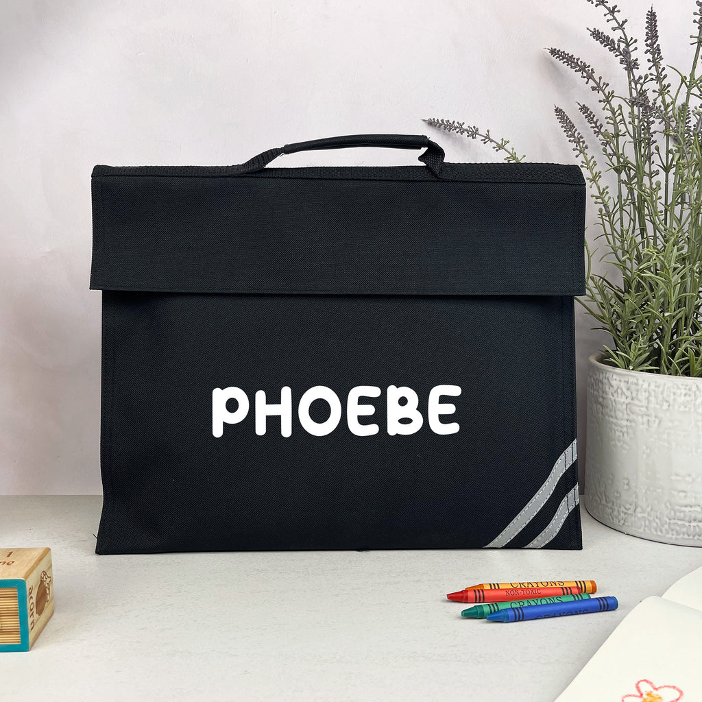 Black bag with 'PHOEBE' printed on it, placed on a surface with crayons and a plant in the background.