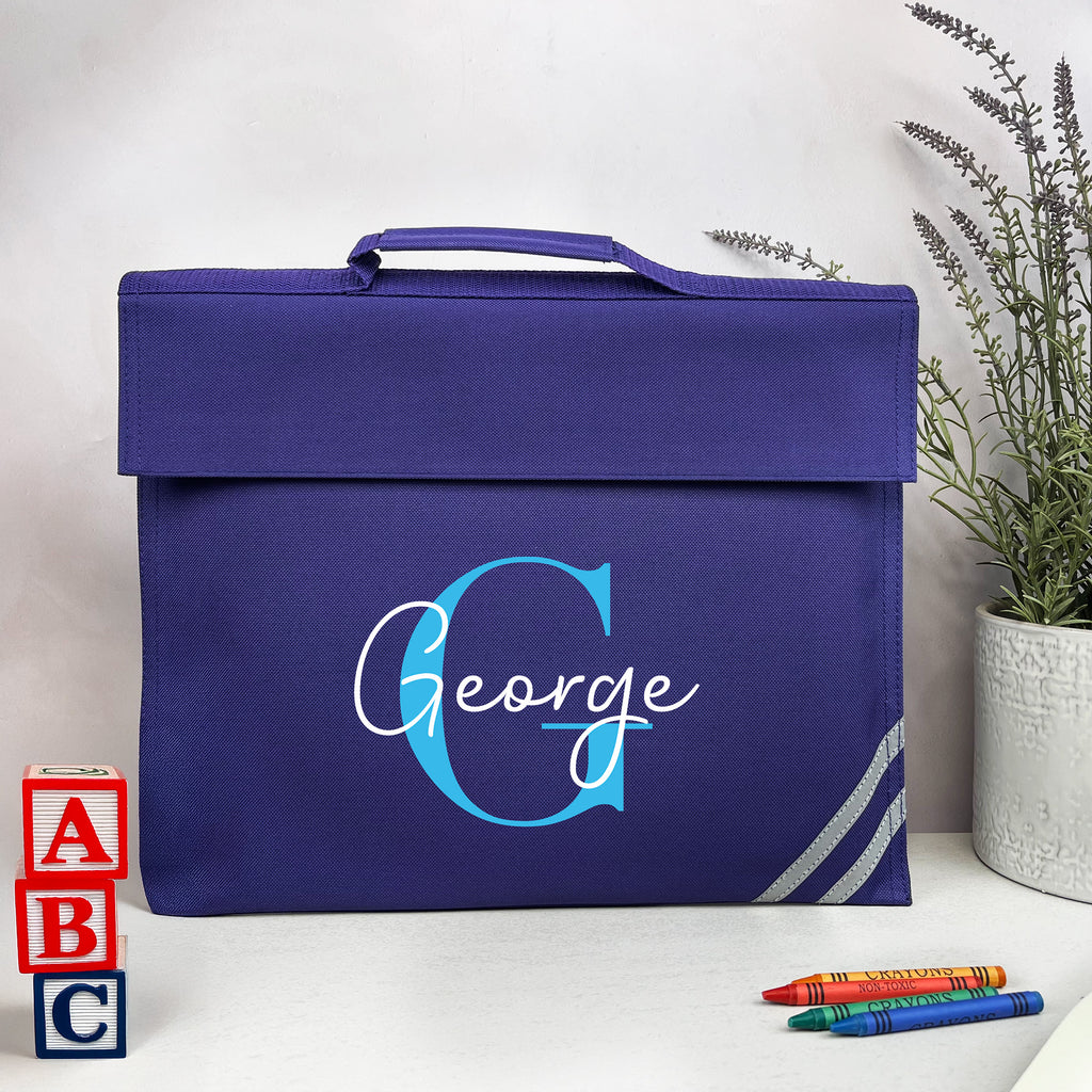 Blue bag with 'George' monogram on a white surface with educational items and a plant.
