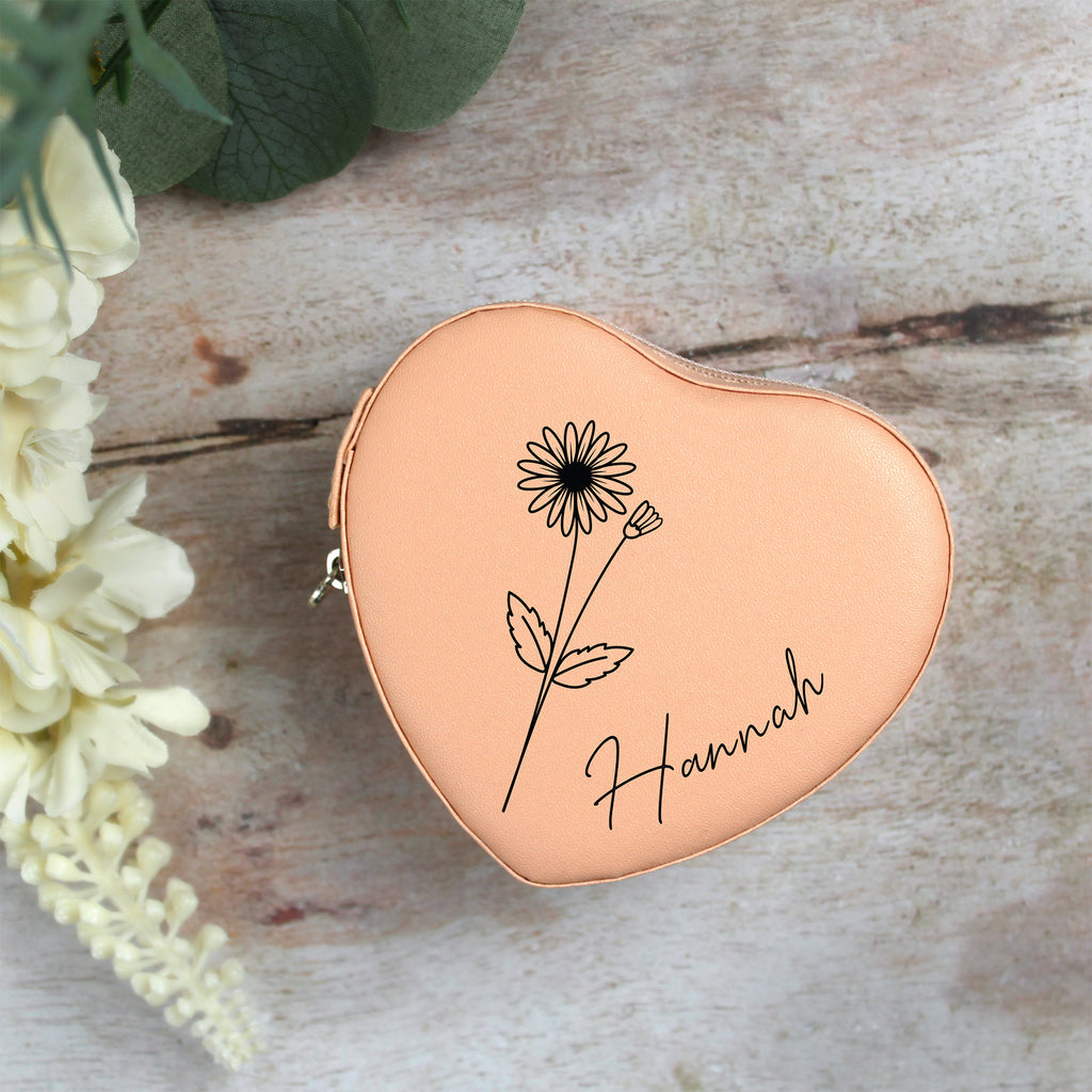 Heart-shaped rose gold pouch with floral design and 'Hannah' engraving on a wooden surface with flowers.