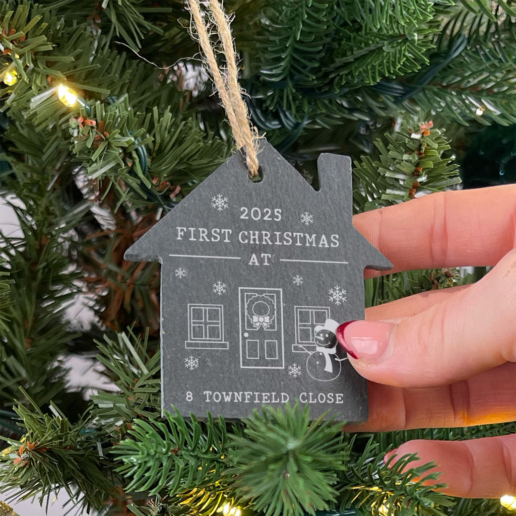 House-shaped ornament with 'First Christmas at' text held against a Christmas tree.