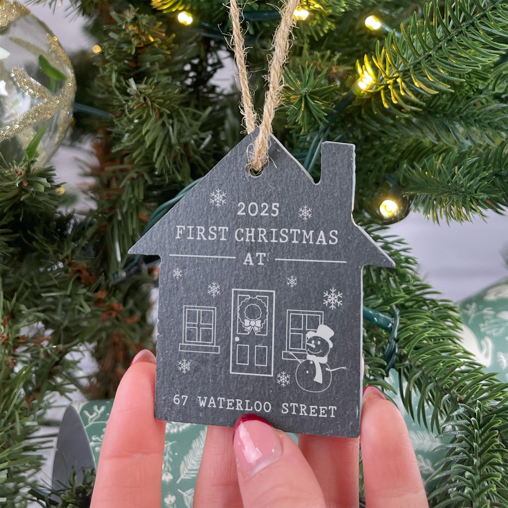House-shaped ornament for first Christmas at a house, held in front of a decorated tree.