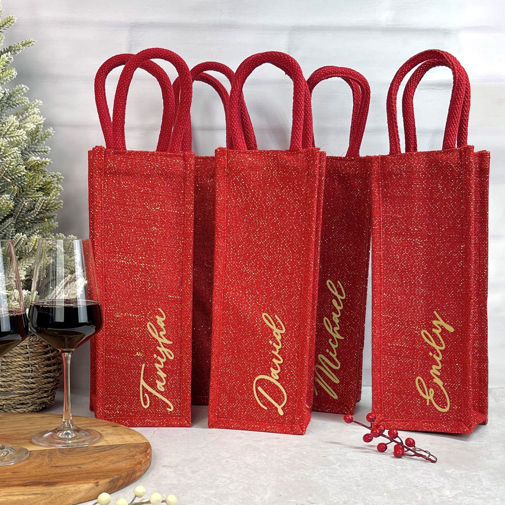 Set of red wine bags with personalized names next to a bottle and glasses on a wooden board.