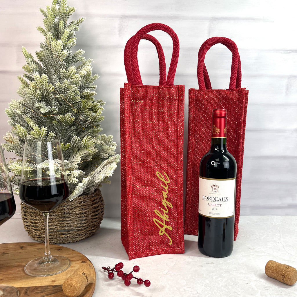 Two red wine bottles in jute bags with a glass of wine and a bottle of wine on a table.