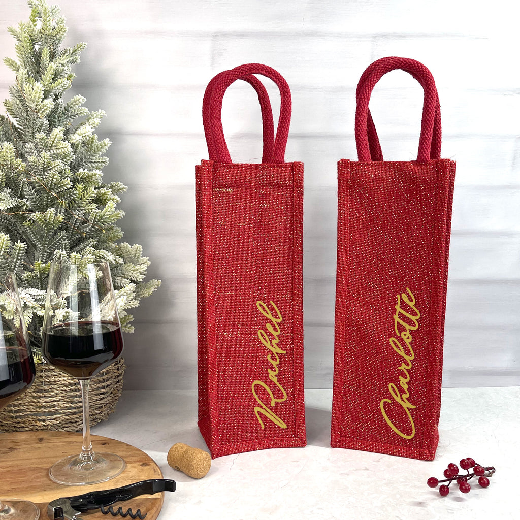 Two red wine bags with gold names next to a glass of wine and a Christmas tree.