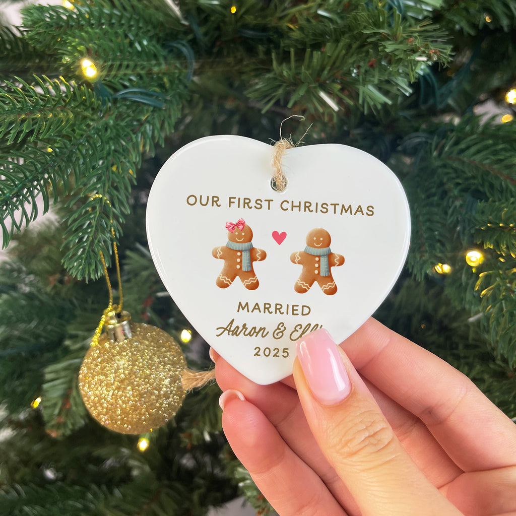 Heart-shaped ornament with 'Our First Christmas Married Aaron & Ellie 2025' text, held in front of a decorated Christmas tree.