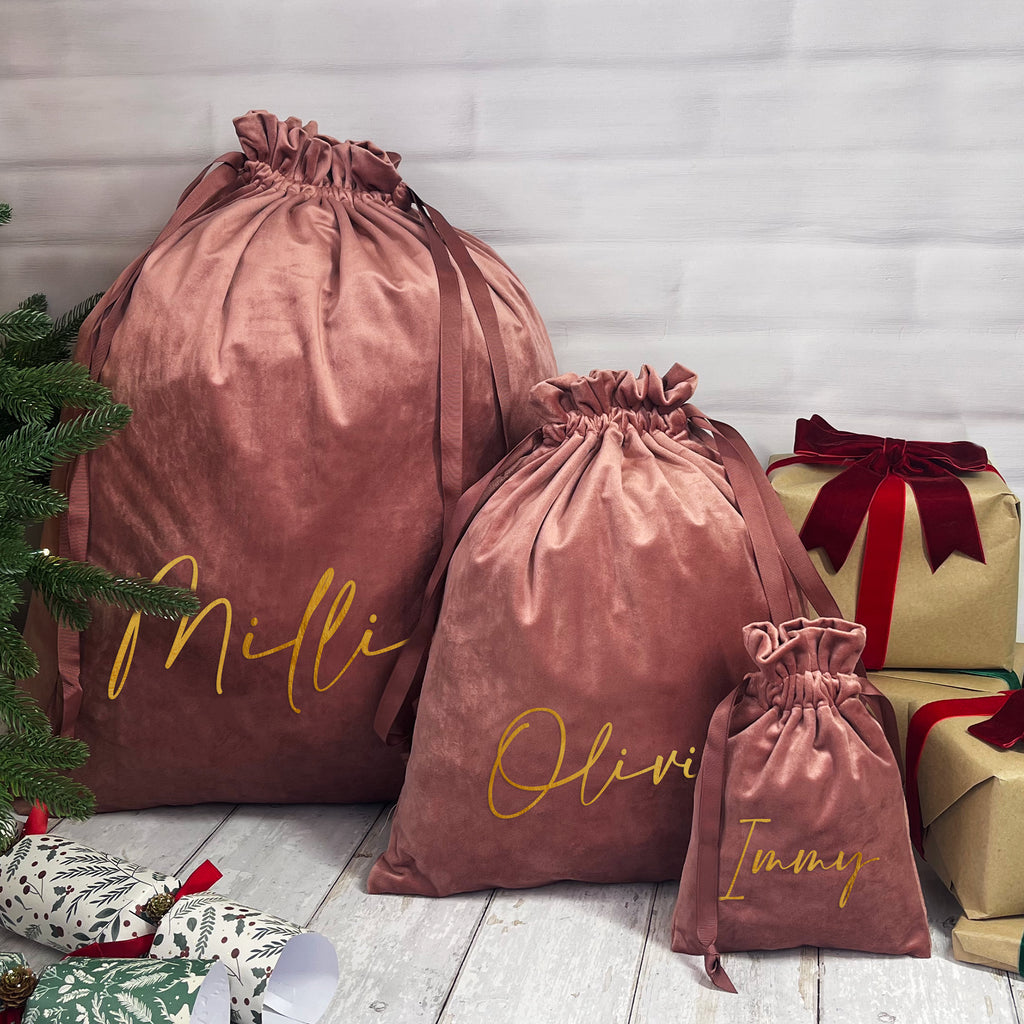 Three drawstring bags labeled 'Mili', 'Olivi', and 'Jimmy' next to Christmas presents on a wooden floor.