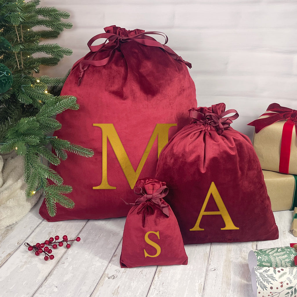 Red velvet sacks with gold letters 'M', 'A', and 'S' next to a Christmas tree and wrapped gifts.