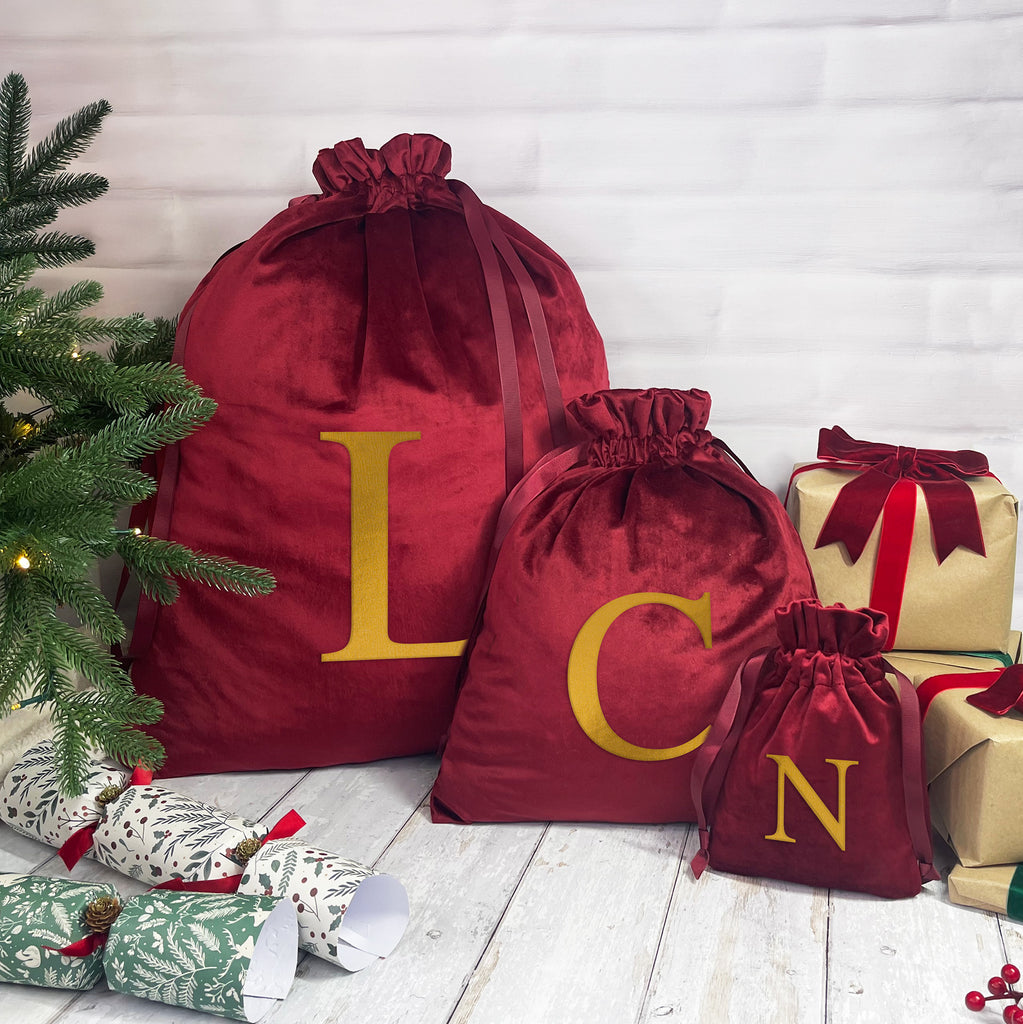 Red drawstring bags with gold letters next to Christmas presents and a tree on a wooden floor.