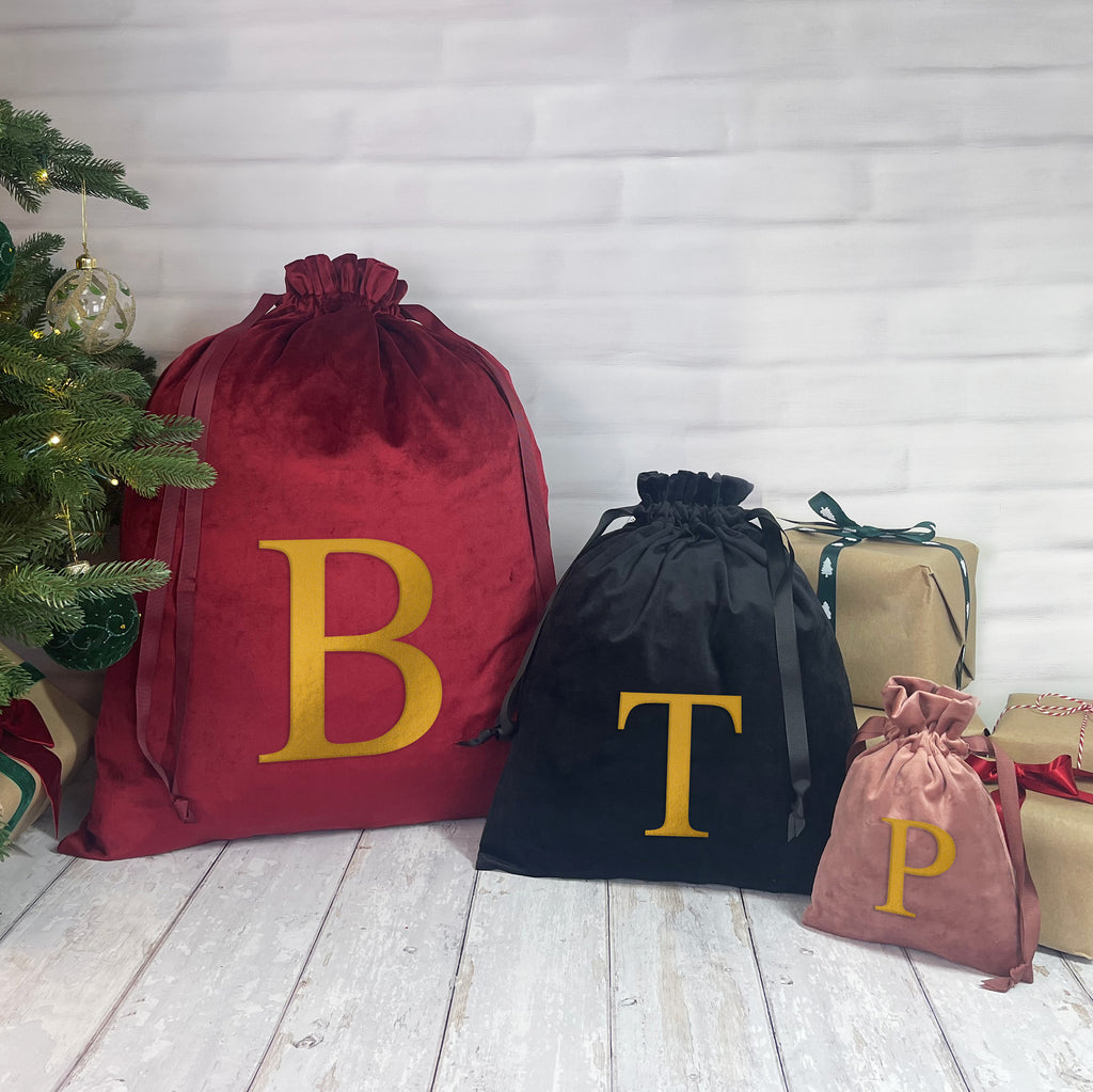 Red, black, and pink drawstring bags with gold letters 'B', 'T', and 'P' next to a Christmas tree.