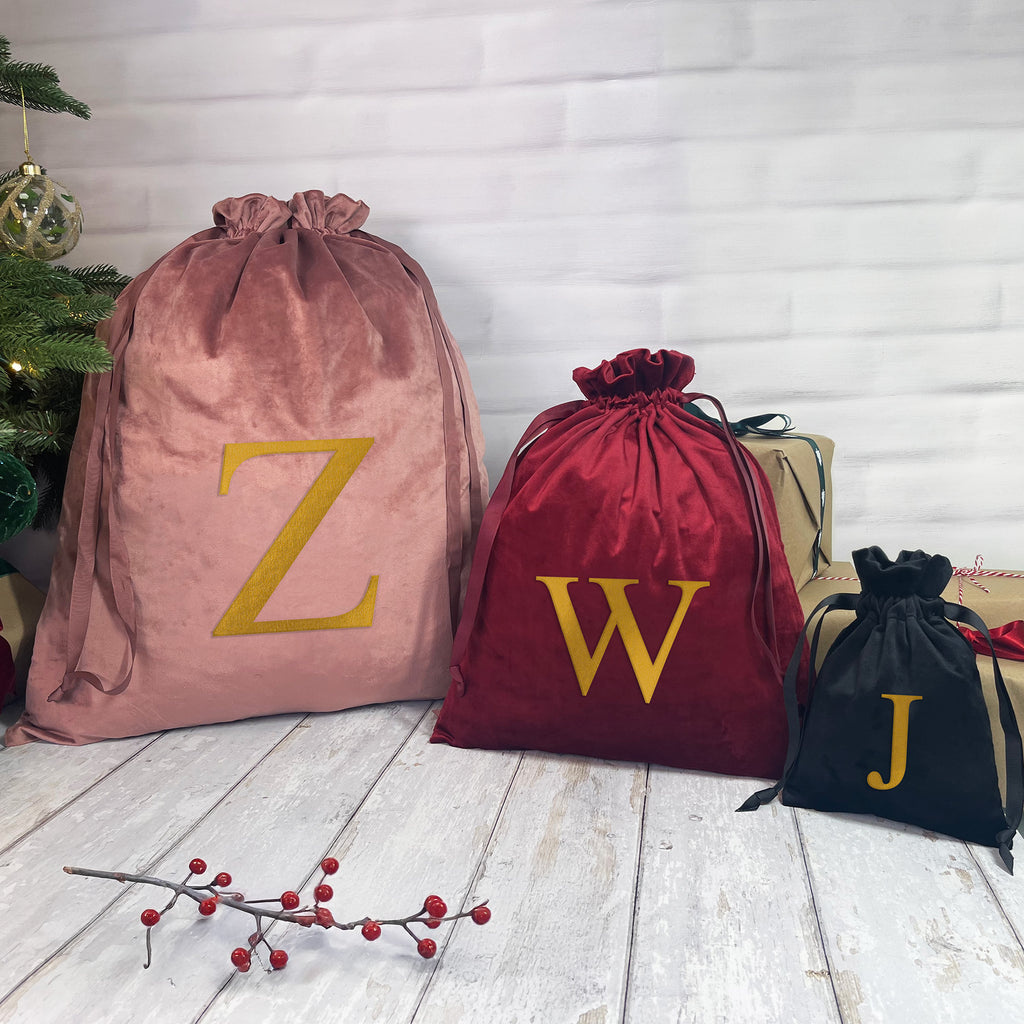 Three drawstring bags with letters 'Z', 'W', and 'J' on a wooden floor with a Christmas tree in the background.