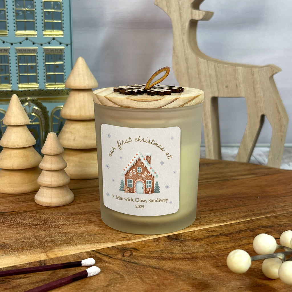 Candle with a gingerbread house design label on a wooden surface with Christmas decorations.