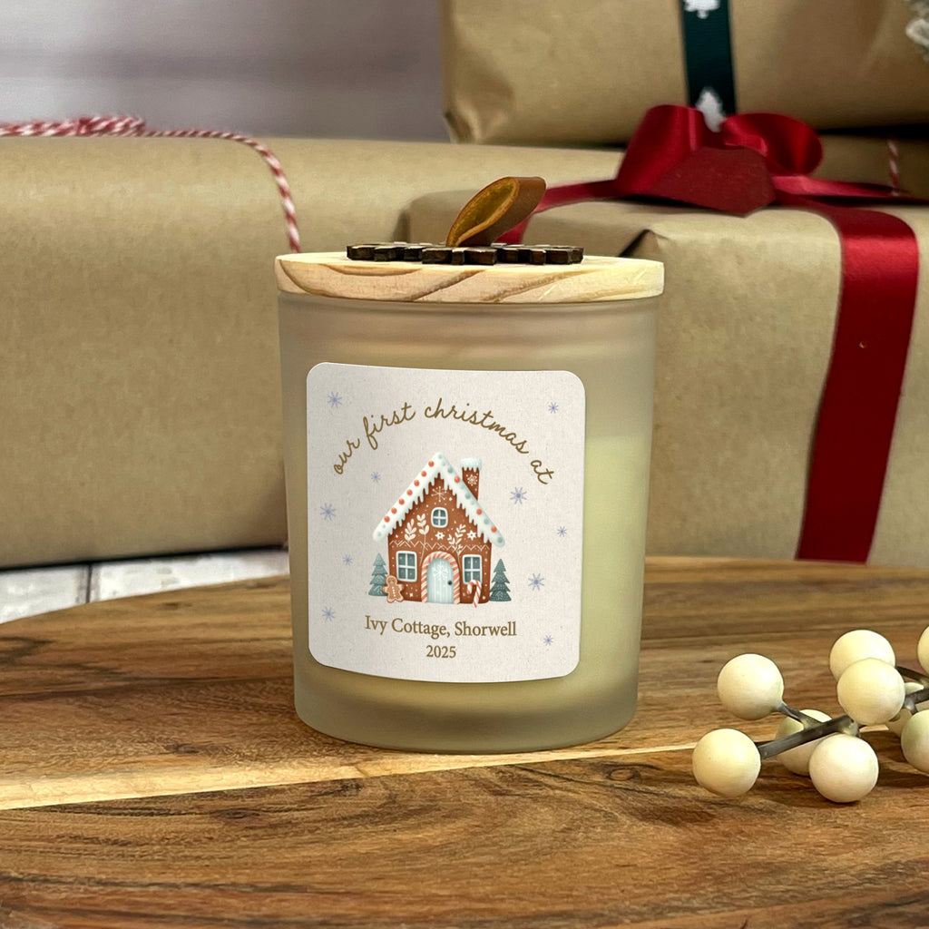 Candle with a gingerbread house design label on a wooden surface with wrapped gifts in the background.
