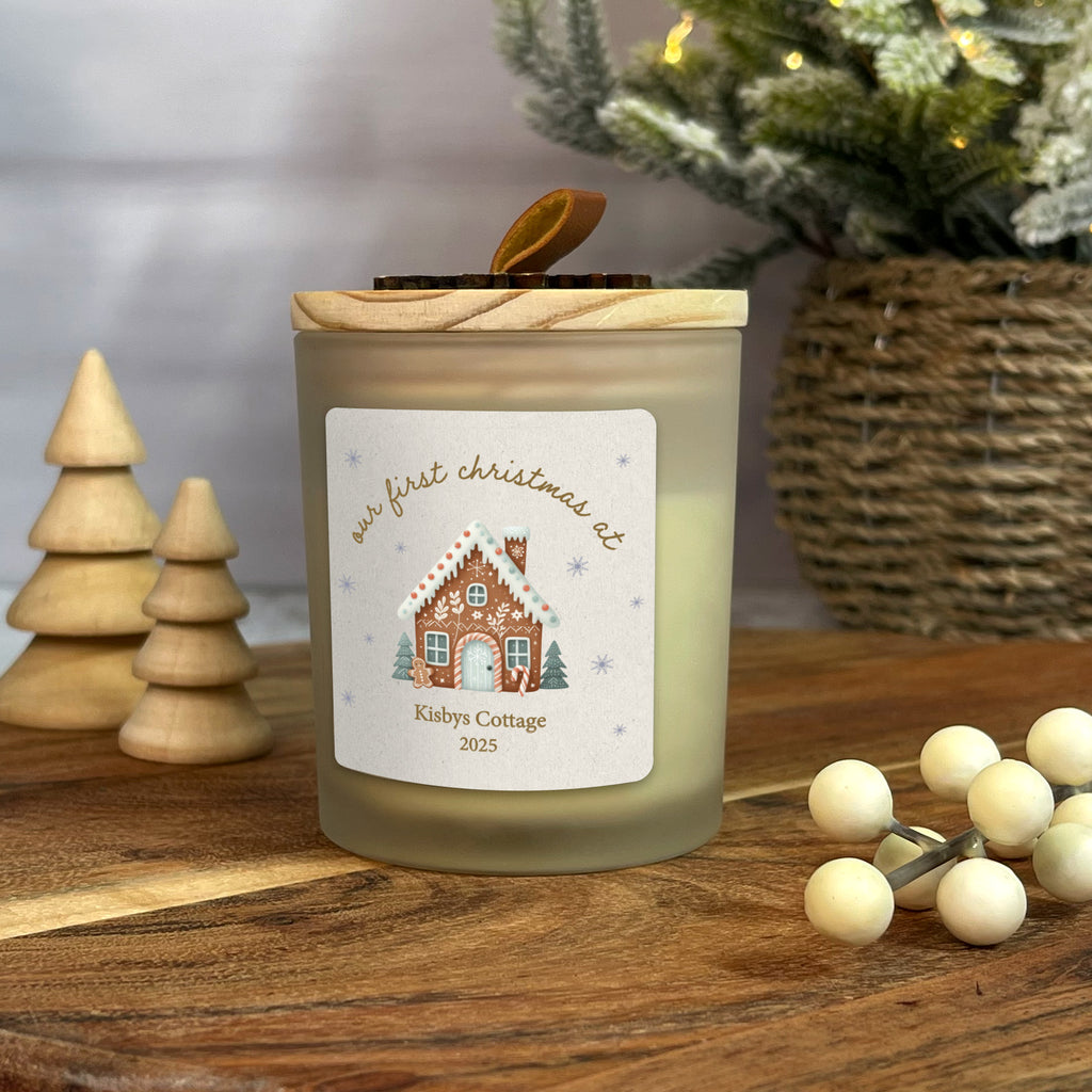 Candle with a gingerbread house design label on a wooden surface with Christmas decorations.