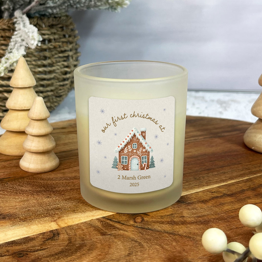 Frosted candle with a gingerbread house design and text on a wooden surface with decorative elements.