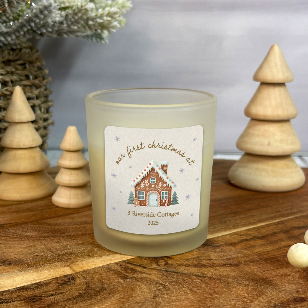 Frosted candle with a Christmas-themed label on a wooden surface with decorative trees.
