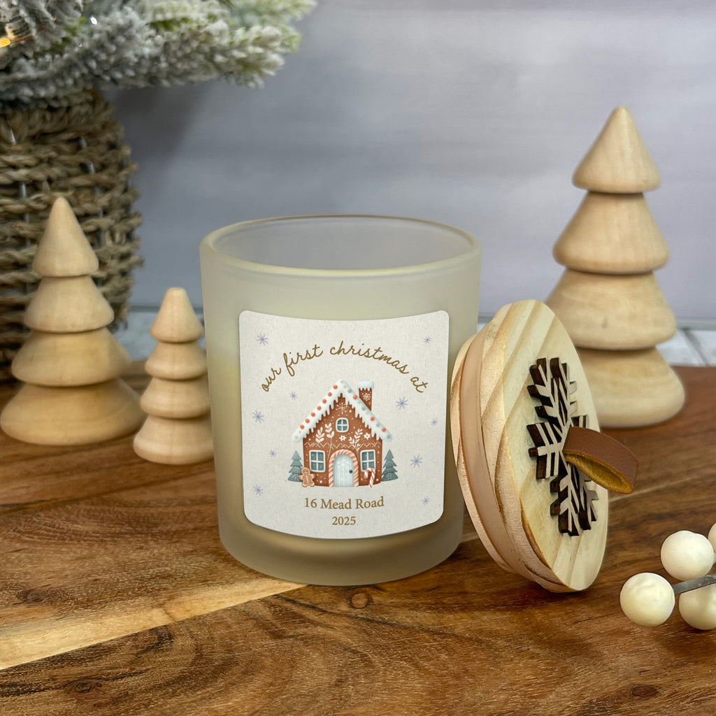 Frosted candle with a gingerbread house design and wooden trees on a wooden surface.
